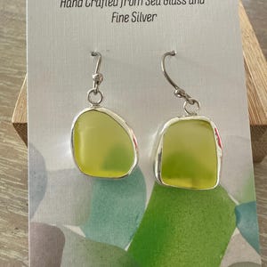 May include: A pair of earrings featuring lime green sea glass encased in silver settings with silver hooks. The display card reads SILVER BUSY BEE and Hand Crafted from Sea Glass and Fine Silver, with a green and blue abstract pattern at the bottom.