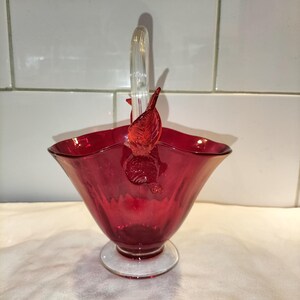 May include: A deep red glass basket featuring a clear glass handle and a clear circular base. The rim of the basket has a wavy, scalloped edge, and the handle is adorned with decorative red glass leaf accents on both sides of the attachment point.