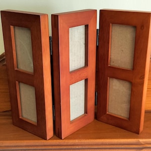 May include: A folding wooden picture frame with three connected panels, each featuring two vertical rectangular openings for photos. The frame has a warm, medium-brown wood finish and is constructed with metal hinges connecting the three sections.