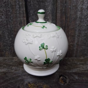 May include: A round white ceramic sugar bowl with a matching lid, featuring embossed and hand-painted green shamrock designs. The bowl has a pedestal base and a small knob handle on the lid, with thin green painted lines accenting the rim and lid.