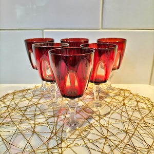 May include: A set of six small glasses with deep red bowls and clear stems. Each glass features a rounded bowl with vertical cut panels that reveal the clear glass beneath the red surface. The glasses are arranged in a cluster on a gold wire mat.