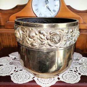 May include: A polished brass planter featuring a wide, embossed band around the upper rim decorated with detailed floral rose motifs. The cylindrical vessel has a smooth, reflective lower body and a dark, open interior, showcasing a metallic finish.