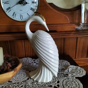 May include: A white ceramic bird figurine with a curved neck and textured, wing-like plumage. The bird features a smooth, glossy finish and a distinct gold-coloured accent on the top of its head, positioned in a downward-facing posture.