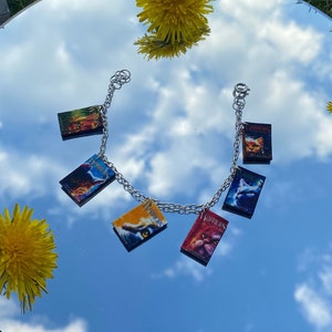May include: A silver chain bracelet with charms featuring covers from the Warriors series books by Erin Hunter. The charms are colorful and feature images of cats.