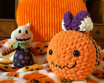 May include: Three crocheted Halloween decorations: a pumpkin, a ghost, and a bee. The pumpkin is orange and has a green stem. The ghost is white with black eyes and a purple scarf. The bee is orange with black stripes and purple wings. The decorations are sitting on a white blanket with a pumpkin pattern.