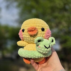 May include: A handmade crochet duck plushie with a yellow body, orange beak and feet, and pink cheeks. The duck is wearing a green frog-shaped accessory. The plushie is soft and cuddly, perfect for children or as a decorative item.