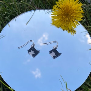 May include: A pair of silver earrings with a black and white star design. The earrings are hanging from a mirror surface.