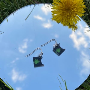 May include: A pair of silver dangle earrings with black and green clover-shaped charms. The earrings are reflected in a mirror.