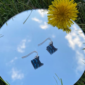 May include: A pair of silver dangle earrings featuring a black and blue outline of the state of Minnesota. The earrings are reflected in a mirror, with a yellow dandelion flower in the background.
