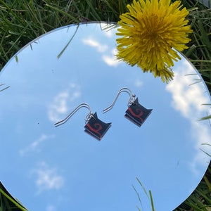 May include: A pair of silver dangle earrings with black and red geometric shapes. The earrings are hanging from a mirror surface.