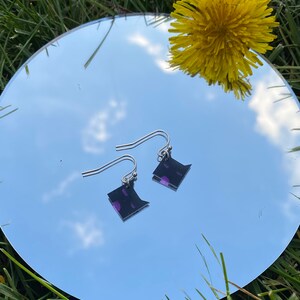 May include: A pair of silver dangle earrings with black and purple square-shaped charms. The earrings are reflected in a mirror.