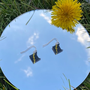 May include: A pair of silver earrings with black and yellow lightning bolt designs. The earrings are hanging from a mirror surface.