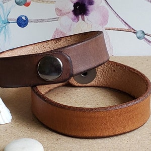 May include: Two leather bracelets in shades of brown. One is a darker brown, and the other is a lighter tan color. Each bracelet has a metal snap closure. The bracelets are displayed on a light brown surface.