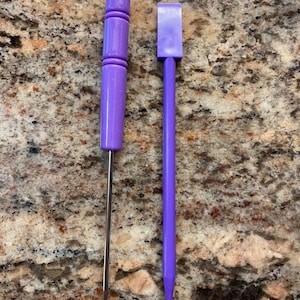 May include: Two purple tools for crafting. One tool has a wooden handle and a metal needle. The other tool has a purple handle and a pointed tip.