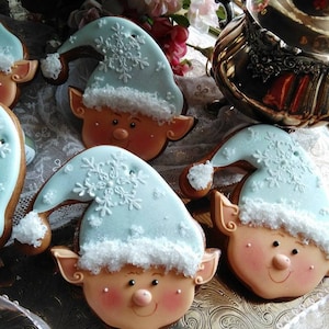 May include: A close-up of four gingerbread elf cookies decorated with white icing and blue frosting. The cookies are on a silver tray with a white lace tablecloth.