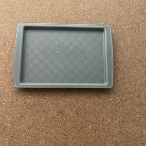 May include: A rectangular, gray plastic tray with rounded corners. The tray has a textured, diagonal pattern on the inside surface. The tray is shown on a brown, textured surface.