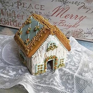 May include: A gingerbread house decorated with white icing, brown gingerbread, and orange and green icing details. The roof is a light blue color with a brown gingerbread trim.