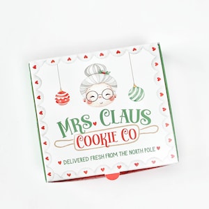 May include: A white box with a green border and red hearts. The box has a cartoon illustration of a woman with glasses and a rolling pin. The text on the box reads "Mrs. Claus Cookie Co. Delivered fresh from the North Pole."