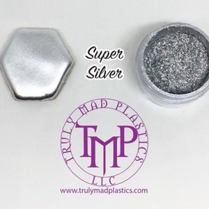 May include: A small jar of silver glitter with the text "Super Silver" printed on a white background. The jar is sitting on a white surface. The jar is labeled "Truly Mad Plastics LLC" and has a website address printed on the bottom.