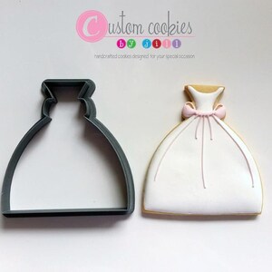 May include: A gray plastic cookie cutter shaped like a wedding dress, next to a decorated sugar cookie in the shape of a wedding dress with a pink bow. The cookie is white with gold icing around the bottom edge.