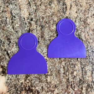 May include: Two purple plastic spatulas with rounded handles and a circular hole in the center of each handle. The spatulas are on a brown and beige speckled countertop.