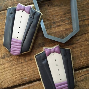 May include: Two decorated sugar cookies shaped like a tuxedo. The cookies are decorated with black icing, white icing, and purple icing. The cookies are on a wooden surface.