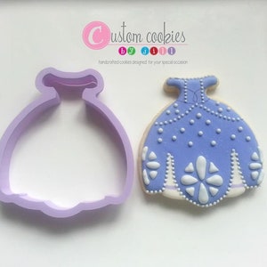 May include: A purple cookie cutter and a decorated purple cookie in the shape of a princess dress. The cookie is decorated with white dots and a white flower design.
