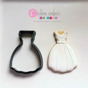 May include: A white cookie decorated to look like a wedding dress, next to a gray plastic cookie cutter in the shape of a wedding dress. The cookie cutter is for making cookies.