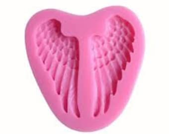 May include: Pink silicone mold shaped like a heart with two angel wings inside. The mold is designed for making cake decorations or other edible crafts.