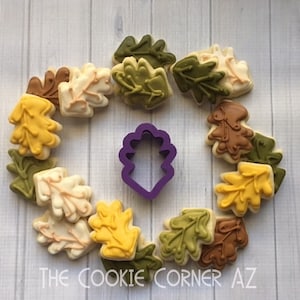 May include: A purple plastic cookie cutter shaped like an oak leaf, surrounded by 13 decorated sugar cookies in various shades of green, brown, and yellow. The cookies are decorated with icing to resemble oak leaves.