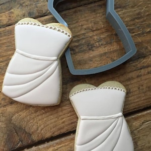 May include: Two white fondant cookies decorated with icing to resemble a strapless wedding dress. A gray plastic cookie cutter in the shape of a strapless wedding dress is also shown.