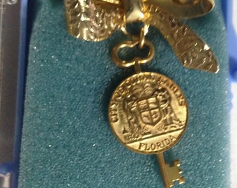 May include: A gold-colored bow-shaped brooch with a key pendant. The pendant features a round medallion with the words "CITY OF MIAMI BEACH FLORIDA" and a coat of arms. The brooch is presented in a blue box.