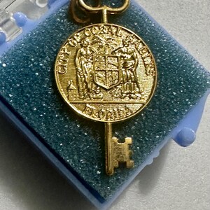 Puede incluir: Charm de llave dorado con un colgante redondo que muestra las palabras "City of Coral Gables, Florida" y un escudo con dos figuras aladas.