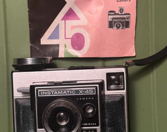 May include: A vintage Kodak Instamatic X-45 camera with a black and silver body, and a matching pink and black instruction manual. The camera has a black lens and the words "Instamatic X-45 Camera" are printed on the front. The manual reads "Kodak Instamatic X-45 Camera".