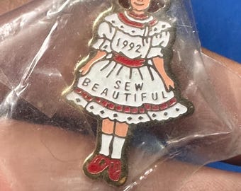 May include: A vintage enamel pin featuring a girl in a white dress with red accents, the text "1992" and "SEW BEAUTIFUL". The pin is in a clear plastic bag.