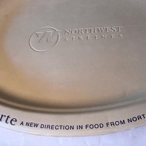May include: Beige oval serving tray with the Northwest Airlines logo and text. The tray has the words "a la carte" and "A NEW DIRECTION IN FOOD FROM NORTHWEST" printed on it.