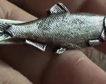 May include: A silver-colored tie clip in the shape of a fish. The fish has detailed scales, fins, and a tail. The clip is designed to attach to a tie, adding a touch of aquatic flair to formal wear.