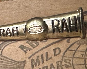 May include: A vintage brass-toned pin with the word "RAH" engraved on it. The pin has a cylindrical shape with a wider, conical section in the middle. The pin is set against a backdrop of a vintage cigar box.