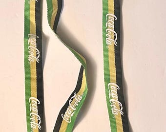 May include: A Coca-Cola branded lanyard with a black, yellow, and green striped pattern. The Coca-Cola logo is printed in white along the length of the lanyard. The lanyard has a white plastic clasp.