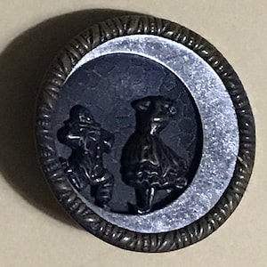 Puede incluir: Un botón negro y plateado con un diseño en relieve de dos figuras en silueta. Las figuras son una mujer con un vestido y un hombre con un sombrero. El botón tiene un borde texturizado.