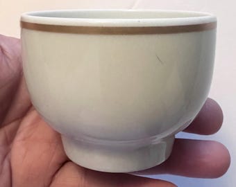 May include: A small, off-white ceramic bowl with a rounded body and a footed base. The rim of the bowl is accented with a thin, brown band. The bowl is held in a person's hand.