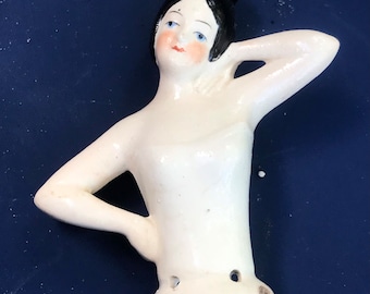 May include: A white porcelain doll with blue eyes and a blue hair bow. The doll is wearing a white dress and has a painted blush on her cheeks.