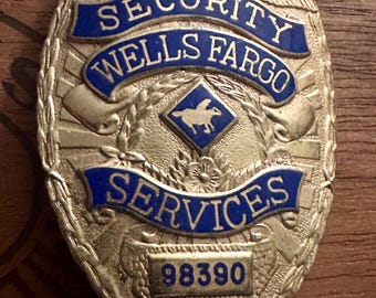 May include: A vintage Wells Fargo Security Services badge. The badge is gold-toned with blue enamel lettering that reads "SECURITY", "WELLS FARGO", and "SERVICES". The badge also features a small emblem and the number "98390".