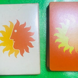 May include: Two playing cards with a sun design. The card on the left is white with a yellow and orange sun design. The card on the right is orange with a yellow sun design.