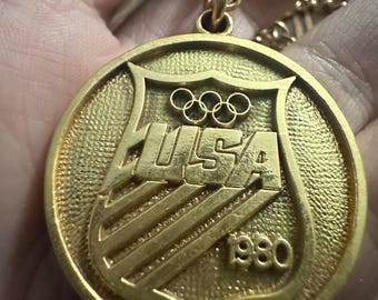 May include: A gold-colored pendant necklace featuring the word "HULSA" in bold letters, the Olympic rings, and the year "1980". The pendant is round with a shield-like design and hangs from a gold chain.