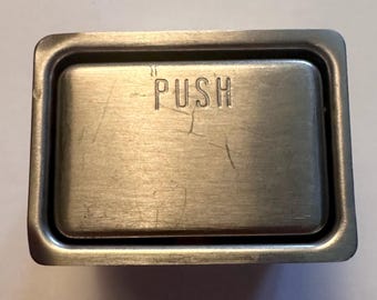 May include: A rectangular, brushed metal push button with the word "PUSH" embossed on the center. The button is slightly recessed within a raised frame. The metal has visible scratches.