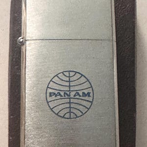Puede incluir: Un encendedor plateado con el logotipo de Pan Am en azul. El encendedor rectangular tiene una tapa con bisagras y se presenta en un estuche marrón oscuro.