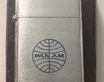 May include: A silver-colored lighter with the Pan Am logo in blue. The lighter is rectangular and has a hinged lid. It is set in a dark brown case.