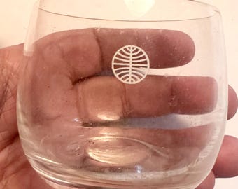 May include: A clear, stemless glass with a rounded base. A white logo is printed on the side. The glass is held in a hand.