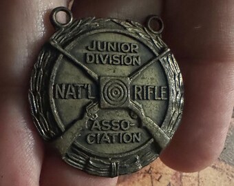 May include: A vintage bronze-colored medal with the text "JUNIOR DIVISION NAT'L RIFLE ASSOCIATION". The medal features crossed rifles, a target, and a laurel wreath border. Two small loops are at the top for attachment.